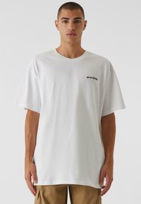 Weißes, oversized T-Shirt aus Baumwolle mit kurzen Ärmeln und Rundhalsausschnitt. Auf der linken Brustseite befindet sich in schwarzem Text "ON1M SENSE."