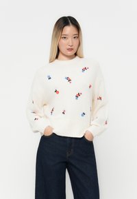 Weißer Strickpullover mit floralen Stickereien in Blau, Rot und Rosa, mit einem gerippten Saum und weiten Ärmeln, kombiniert mit dunkelblauen Jeans.