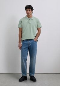 Nuori mies, joka on pukeutunut vaaleanvihreään polo-paitaan, sinisiin farkkuihin ja mustiin kenkiin, seisoo yksi käsi taskussa tasasävyistä taustaa vasten.