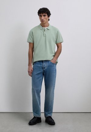 Jonge man in een lichtgroene polo, blauwe spijkerbroek en zwarte schoenen, staand met één hand in zijn zak tegen een effen achtergrond.