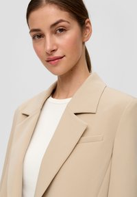 Beige Blazer mit strukturiertem Revers und einer einzelnen Tasche. Darunter befindet sich ein weißes, geripptes Shirt. Glatter Stoff mit maßgeschneidertem Schnitt.