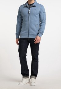 Blå zip-up sweatshirt med hög krage, två framfickor, ribbade ärmar och en slät textur, tillsammans med mörka jeans och vita sneakers.