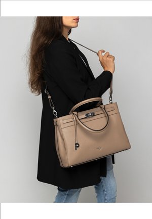 Woman wearing black coat and blue jeans holds taupe leather handbag with shoulder strap and top handles against gray background.