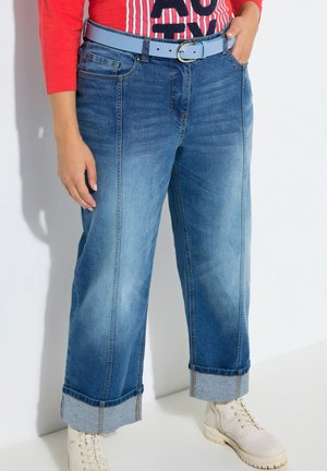 Modré džíny s širokými nohavicemi do poloviny výšky, ohrnuté lemy, světle modrý pásek se stříbrnou přezkou, kombinované s bílými kozačkami na šněrování a červenou košilí zastrčenou dovnitř.