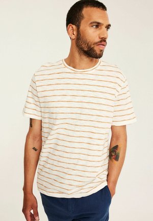 Man met kort haar en baard, draagt een wit shirt met korte mouwen met dunne oranje horizontale strepen en blauwe broek, kijkt naar rechts.