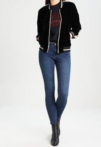Giacca bomber in velluto nero con polsini a righe, indossata sopra una maglietta nera con grafica, abbinata a jeans skinny blu e stivaletti neri.