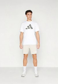 T-shirt branca com um logótipo adidas camuflado, combinada com calções bege que exibem um logótipo de texto adidas em branco. O modelo usa meias e sapatilhas brancas.