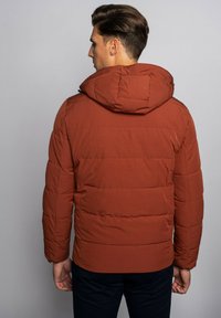 Bruine pufferjas met capuchon, voorzien van een doorgestikt ontwerp en elastische manchetten. De achterzijde toont een gladde textuur en minimale details.