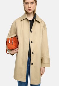 Beżowy trencz z dużym kołnierzem, z przednimi guzikami i bocznymi kieszeniami, w komplecie z małą pomarańczową torbą z zamszu z metalowymi zamkami błyskawicznymi.