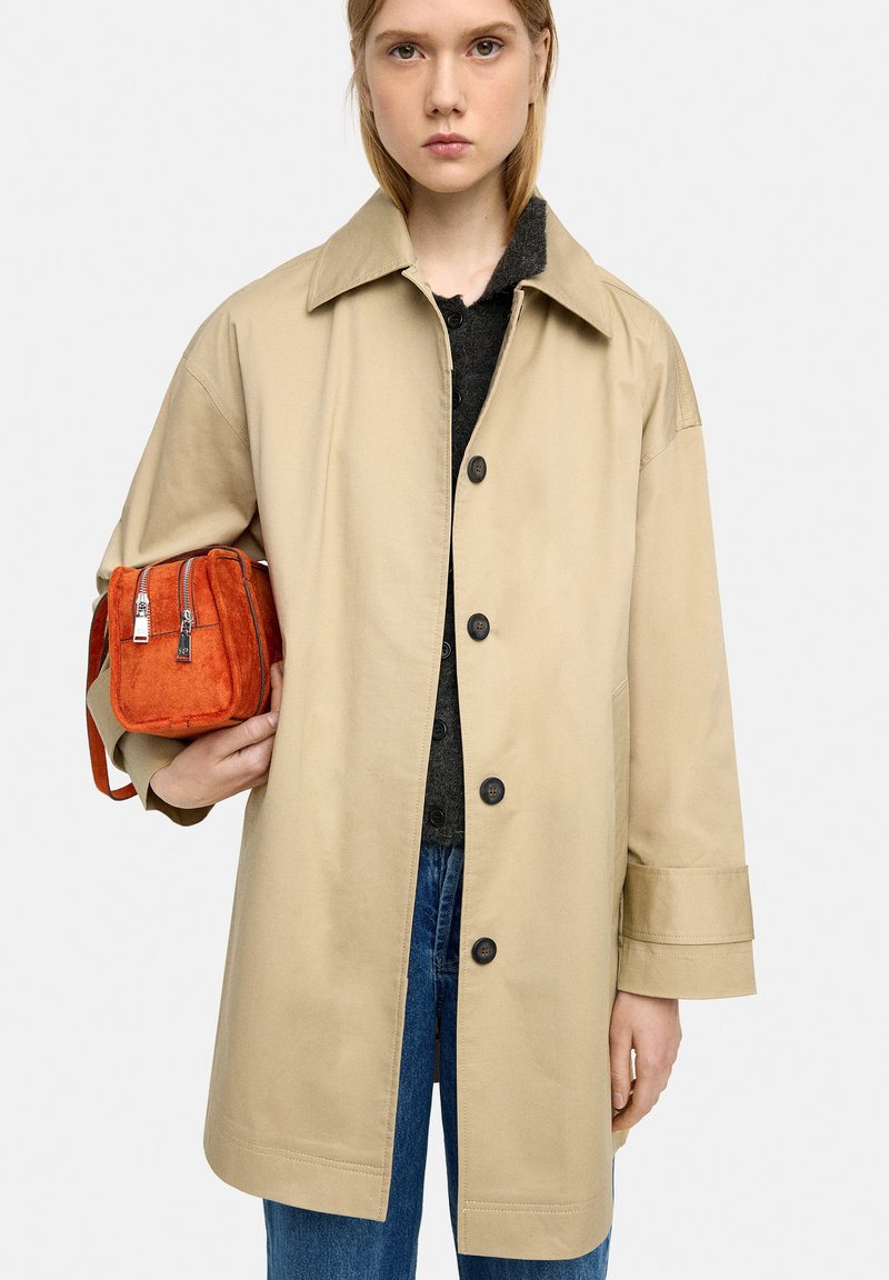 Beżowy trencz z dużym kołnierzem, z przednimi guzikami i bocznymi kieszeniami, w komplecie z małą pomarańczową torbą z zamszu z metalowymi zamkami błyskawicznymi.