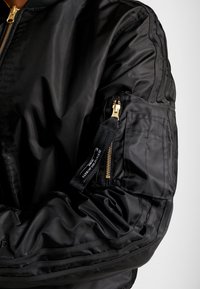 Svart bomberjacka med blank finish, blixtlåsficka på ärmen och guldfärgade detaljer. Har strukturerade mönster på tyget.