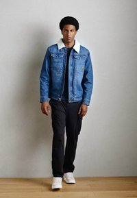 Denimjacke mit weißem Schafwollkragen, zwei Brusttaschen, kombiniert mit schwarzen Cargo-Hosen und weißen Sneakers. Glatte Stofftextur.