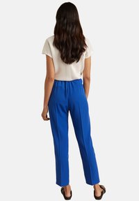 Pantalon bleu avec une coupe décontractée et des plis, fabriqué dans un tissu lisse, associé à un haut blanc à manches courtes et des sandales noires à enfiler.