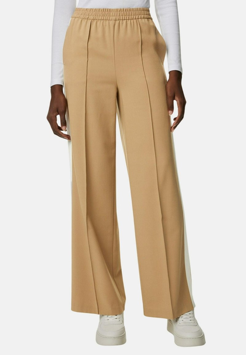 Personne portant un pantalon beige à jambes larges avec une taille élastique, assorti à des baskets blanches et un haut blanc à manches longues.