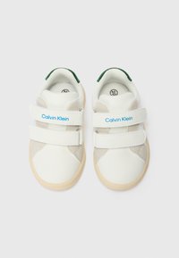 Vita barn sneakers med en slät yta, utrustade med två kardborreband, ljusgrå detaljer och en beige gummisula. "Calvin Klein"-logotypen är synlig.