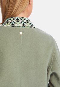 Grüne Wollmischung Jacke mit einem schwarzen und gemusterten Kragen, ausgestattet mit einem goldenen Akzentknopf auf der Rückseite und einer glatten Textur.