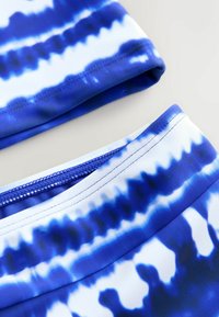 Sinine ja valge tihedusega ujumisriiete ülaosa ja alaosa, mis on valmistatud siledast ja venivast kangast, sisaldades tugevdatud õmblust ja nähtamatut riistvara.