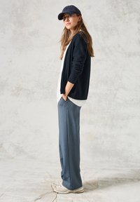 Mörkblå cardigan över en vit skjorta, i kombination med ljusblå byxor och beige sneakers. Personen bär en mörkblå keps, med händerna i fickorna.