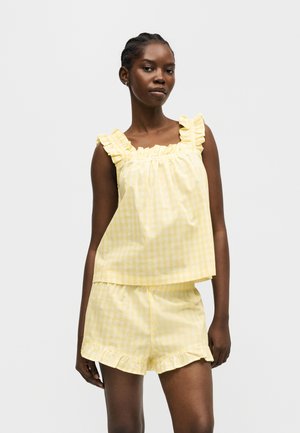 Žena má na sebe žltý kockovaný tielko bez rukávov s volánikovými ramienkami a krátke nohavice s volánikmi na spodnom okraji na jednofarebnom pozadí.