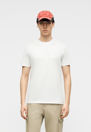 Junger Mann mit roter Baseballkappe, weißem kurzärmeligem T-Shirt und beigen Cargohosen vor einem einfarbigen hellen Hintergrund.