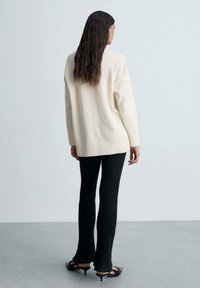 Pulover oversized crem cu tiv ribetat, asortat cu pantaloni evazați negri cu textură ribetată și pantofi deschisi la vârf negri, având o textură netedă.