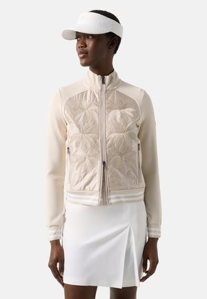 Kobieta ubrana w beżową kurtkę na zamek z haftem liścia palmy, białą spódnicę z rozcięciem z przodu oraz biały daszek, lekko zwrócona w lewo.