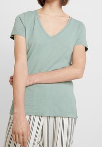 Ljusgrön t-shirt med kort ärm och V-ringning. Tillverkad av mjukt material och har en avslappnad passform. Modellen poserar med armarna i kors och bär randiga byxor.