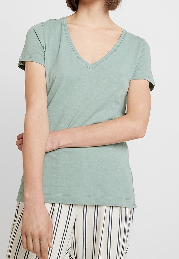 Ljusgrön t-shirt med kort ärm och V-ringning. Tillverkad av mjukt material och har en avslappnad passform. Modellen poserar med armarna i kors och bär randiga byxor.