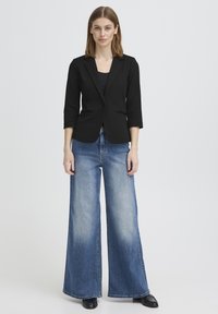 Nainen seisoo eteenpäin katsovana pukeutuneena mustaan bleiseriin, mustaan topiin, leveälahkeisiin haalistuneisiin sinisiin farkkuihin ja mustiin kenkiin, vaaleaa taustaa vasten.