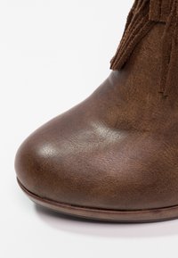 Bout de botte en cuir marron avec une texture lisse et une tige en daim à franges visible sur fond blanc.
