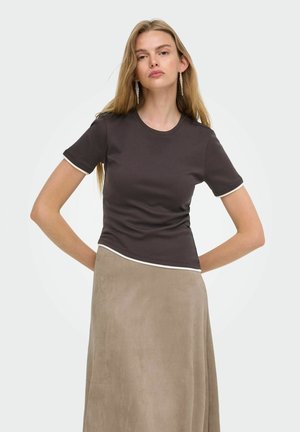 Blondynka w ciemnobrązowym topie z krótkim rękawem z białą lamówką i jasnobeżową długą spódnicą, stoi z rękami za plecami.