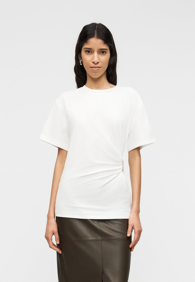 Γυναίκα με μακριά σκούρα μαλλιά, που φορεί λευκή μπλούζα με κοντά μανίκια και πτυχωτό σχέδιο στο πλάι, και σκουρόχρωμη δερμάτινη φούστα, στέκεται μπροστά σε απλό φωτεινό φόντο.