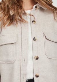 Lys grå uldjakke med to brystlommer, sorte og brune knapper samt en blød tekstur. Hvid ribbet skjorte synlig nedenunder.