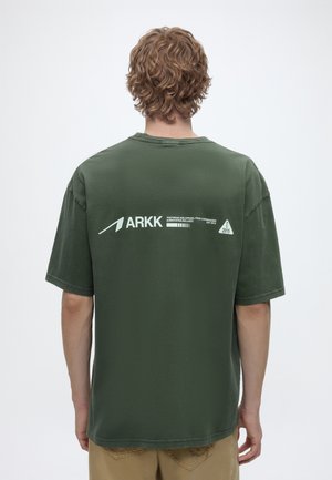 Personne aux cheveux bouclés portant un T-shirt vert olive avec le logo et le texte blanc "ARKK" sur le haut du dos.