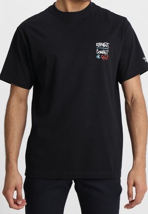 Homme portant un t-shirt noir à manches courtes avec un petit texte coloré sur le côté gauche de la poitrine indiquant "Element Commit or Quit".