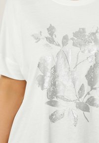 Hvid bomulds T-shirt med et sølvfarvet blomstermotiv med blanke detaljer og en struktureret finish. Løs pasform med korte ærmer.