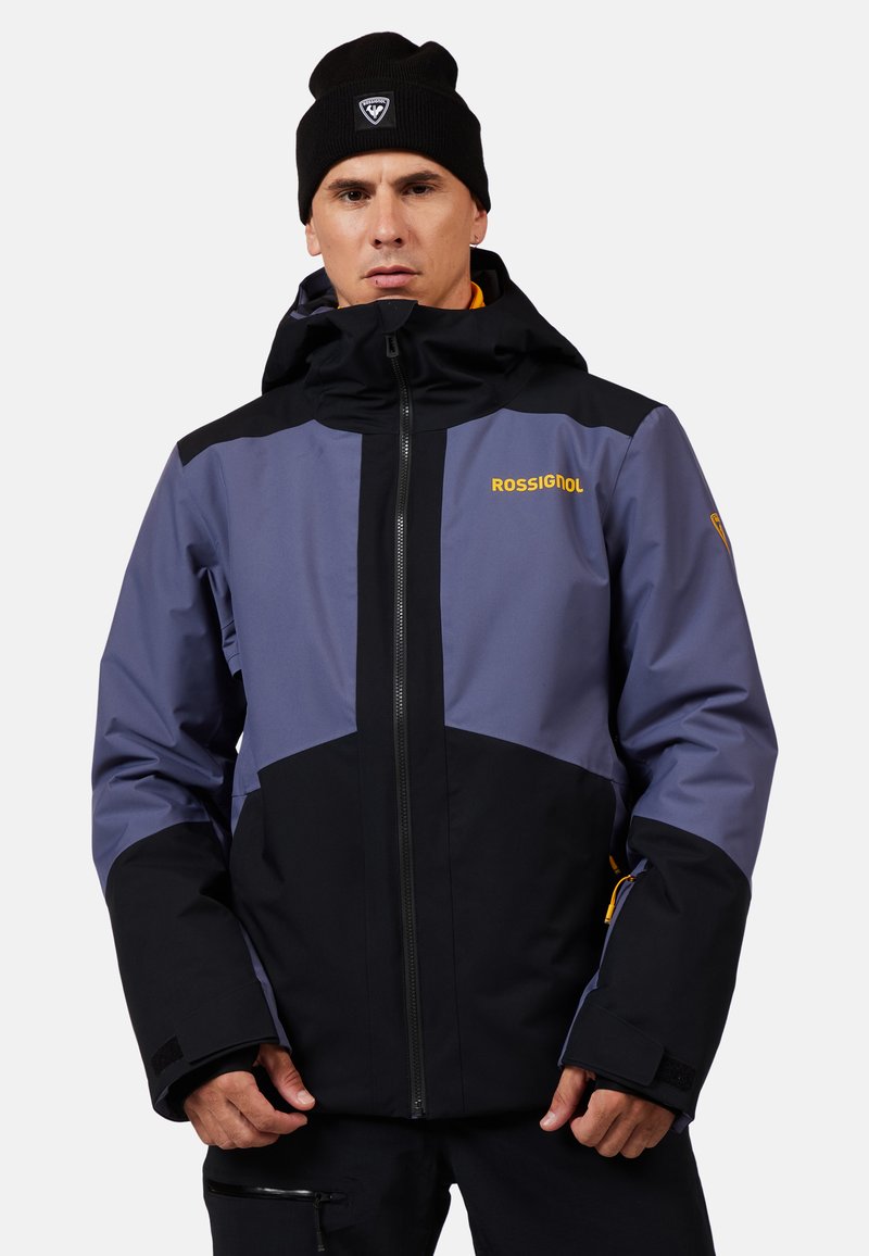 Herrenjacke von Rossignol mit einem Farbblockdesign in Dunkelblau, Schwarz und gelben Akzenten. Hergestellt aus wasserdichtem Material, mit Kapuze und verstellbaren Manschetten.