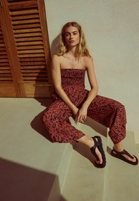 Röd och svart mönstrad strapless jumpsuit med smockad bodice och lösa vida ben, i kombination med svarta sandaler på en beige trappa.