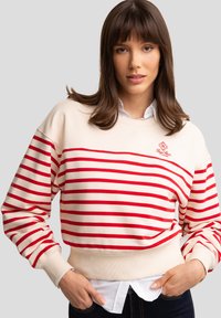 Sudadera crema con rayas horizontales rojas, dobladillo acanalado y mangas ajustadas. Presenta un pequeño logo en el pecho. Lleva puesta sobre una camisa blanca de cuello.