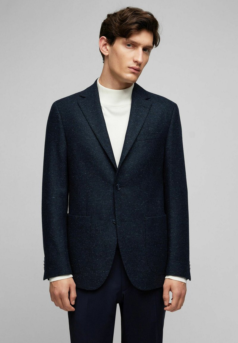 Marineblauer Wollblazer mit strukturiertem Finish, zwei Fronttaschen, Reverskragen und einem einzelnen Knopfverschluss, kombiniert mit einem weißen Rollkragenpullover.