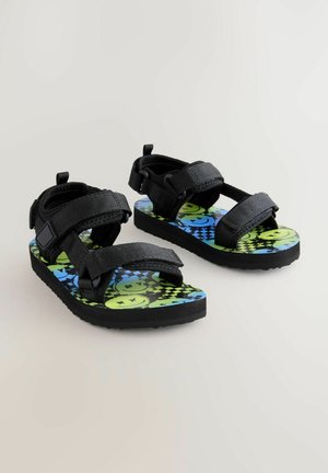 Sandales noires ajustables avec des sangles en tissu et une semelle à motifs présentant des visages souriants verts et bleus sur un fond noir.