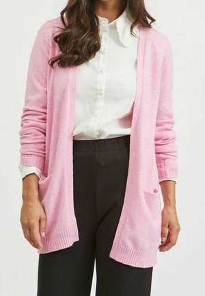 Vrouw draagt een lichtroze open cardigan over een wit gebuttond overhemd met een zwarte broek, tegen een effen achtergrond.