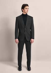 Maßgeschneiderter dunkelgrauer Anzug mit Reverskragen, einem Knopf und vorderen Taschen, kombiniert mit einem schwarzen Rollkragenpullover und schwarzen Lederschuhen.