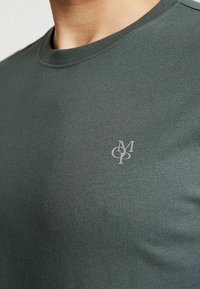 Grünes T-Shirt mit kurzen Ärmeln aus glattem Stoff. Mit einem runden Ausschnitt und einem dezenten Logo in einer helleren Farbe auf der Brust.
