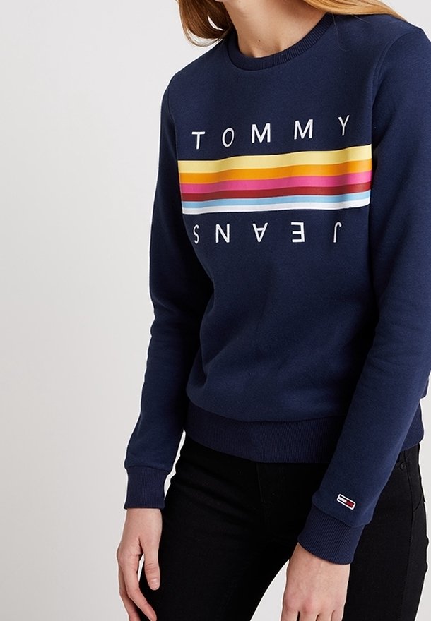 Person som bär en marinblå sweatshirt med horisontella regnbågsränder och texten "TOMMY JEANS" på bröstet samt logotyp på handleden.