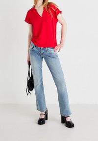 Punainen v-kaula-aukollinen paita yhdistettynä vaalean sinisiin suoralahkeisiin farkkuihin. Asun kruunaavat mustat korotettu alustakengät. Yksinkertainen musta käsilaukku kädessä.