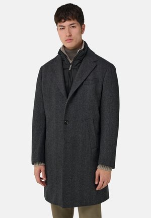 Cappotto grigio in lana con rever a intaglio, chiusura con un solo bottone e due tasche laterali. Indossato sopra una giacca imbottita nera con collo alto.
