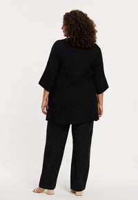 Zwarte tuniek van een gestructureerde stof, met driekwartmouwen en een gebogen zoom, gecombineerd met zwarte wijde broek en sandaaltjes met banden.