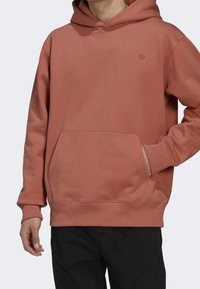 Person som bär en rostfärgad huvtröja med känguruficka och subtil logotyp på bröstet, i kombination med svarta byxor.