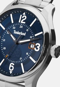 Montre bracelet en métal avec un cadran bleu marine, des index blancs et des aiguilles argentées ; affiche la date à 15 heures et comporte le logo Timberland.